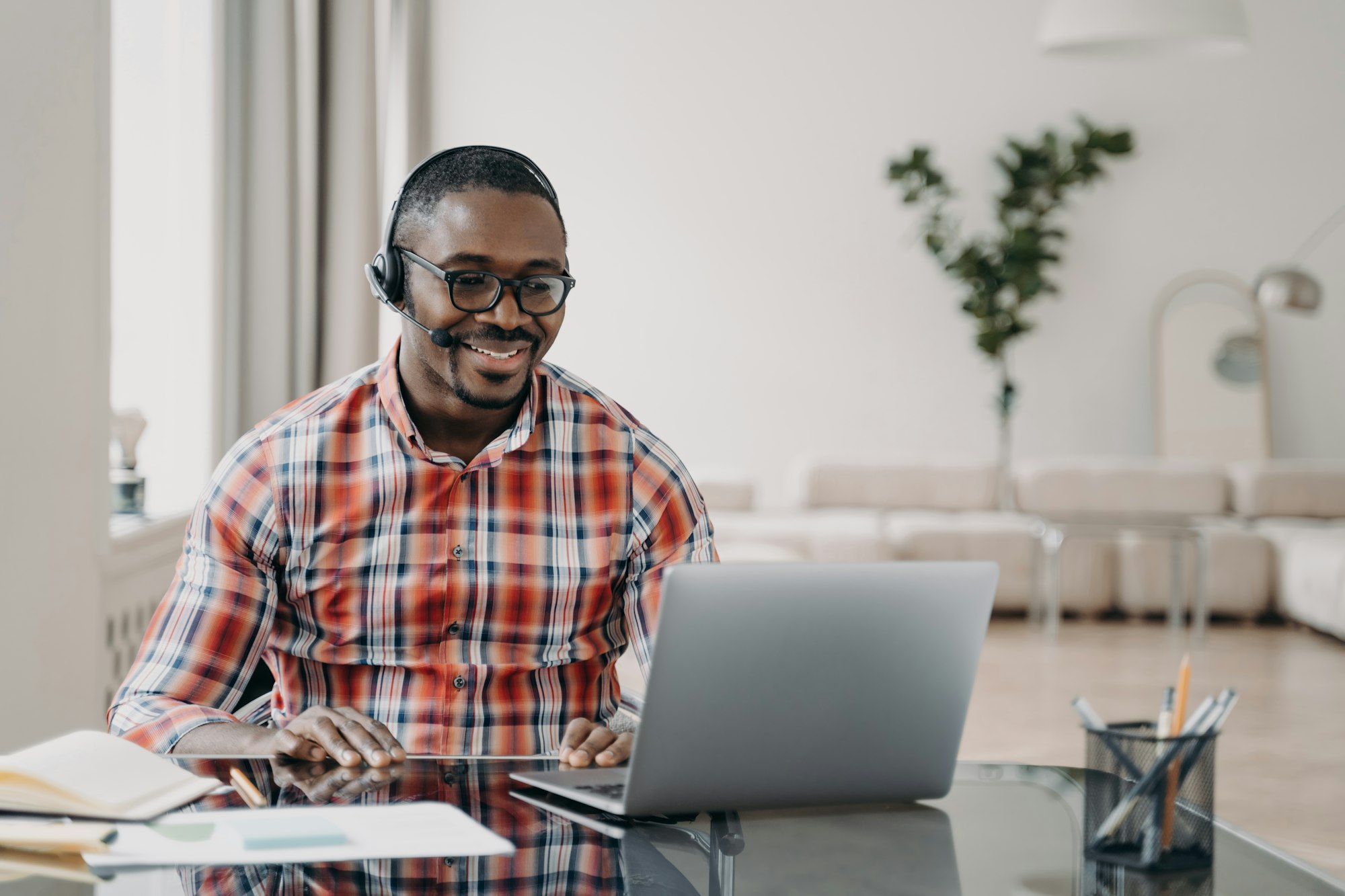 Smiling man in casual plaid shirt working on laptop with headset.